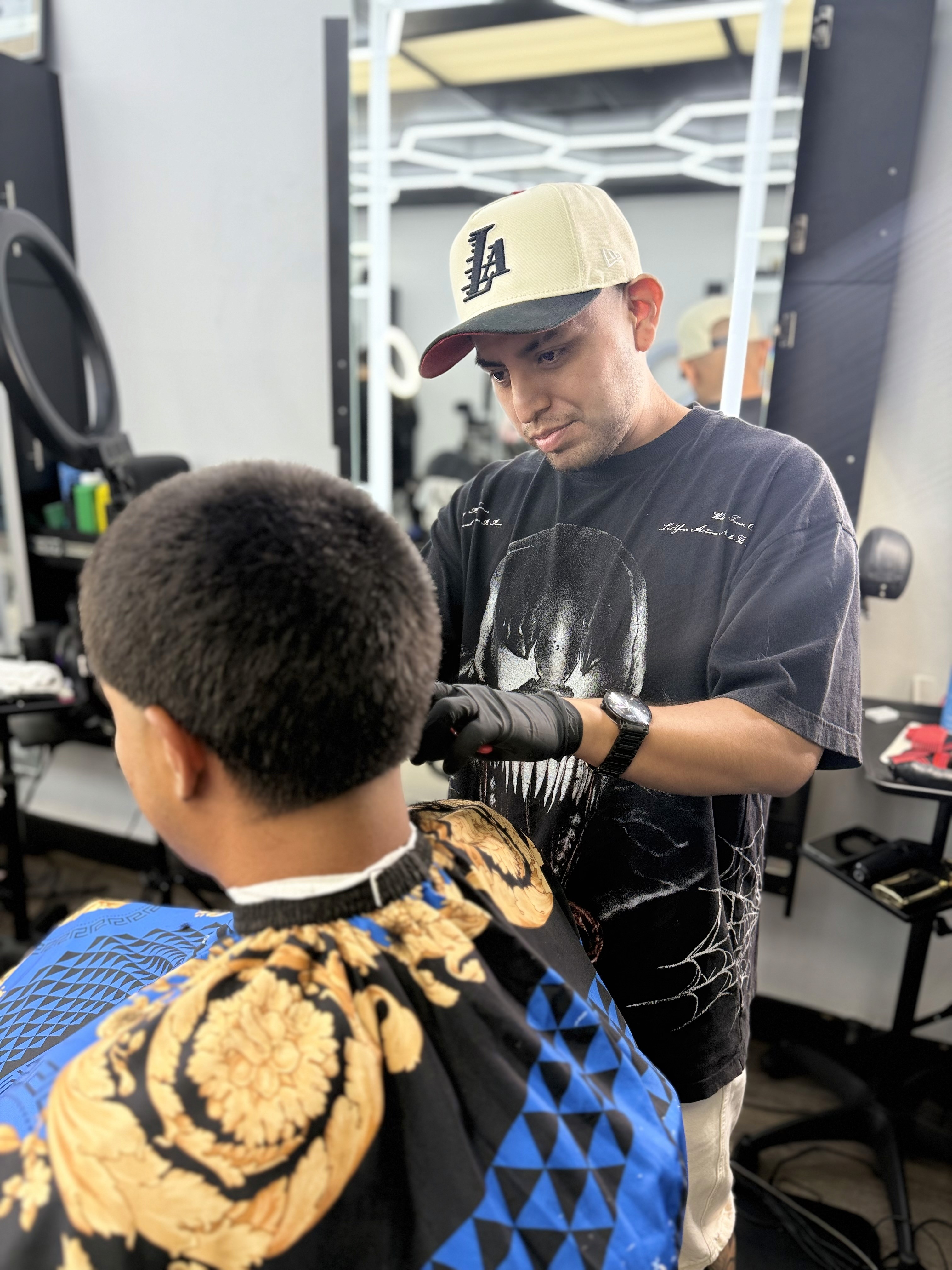 A barber shapes a client’s haircut using clippers near a bright ring light in barbershop. 