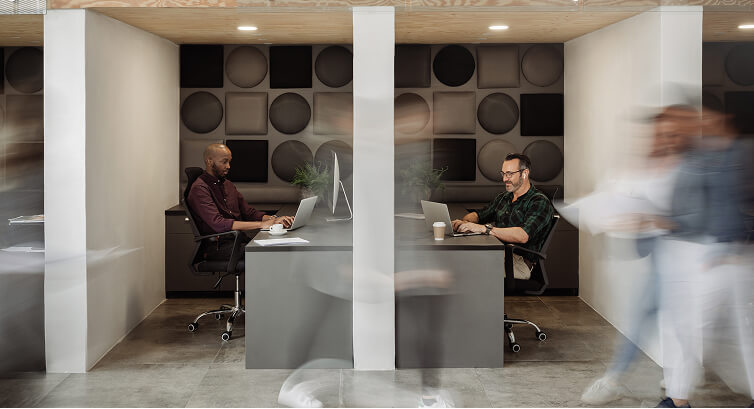 Modern office workspace with white pod-style meeting areas where business professionals are working, with one person walking past in motion blur, featuring contemporary circular wall art