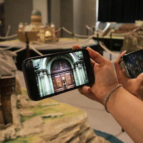 兩人手持智能手機拍攝建築模型的影像，重點聚焦於一個精細的建築立面模型。