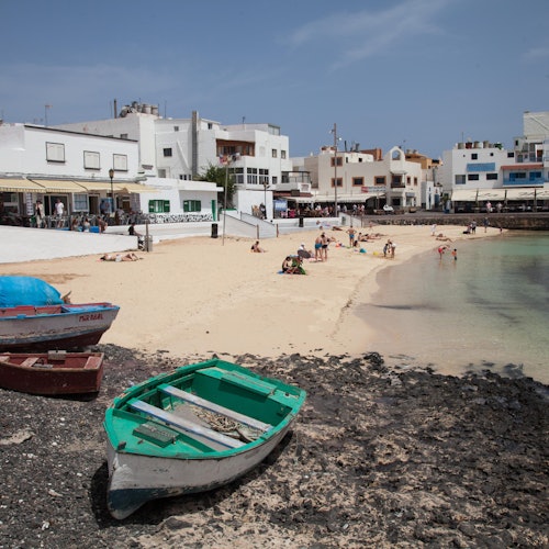 Small boats on rocky shore, people relaxing on sandy beach, and white buildings in the background under a blue sky.