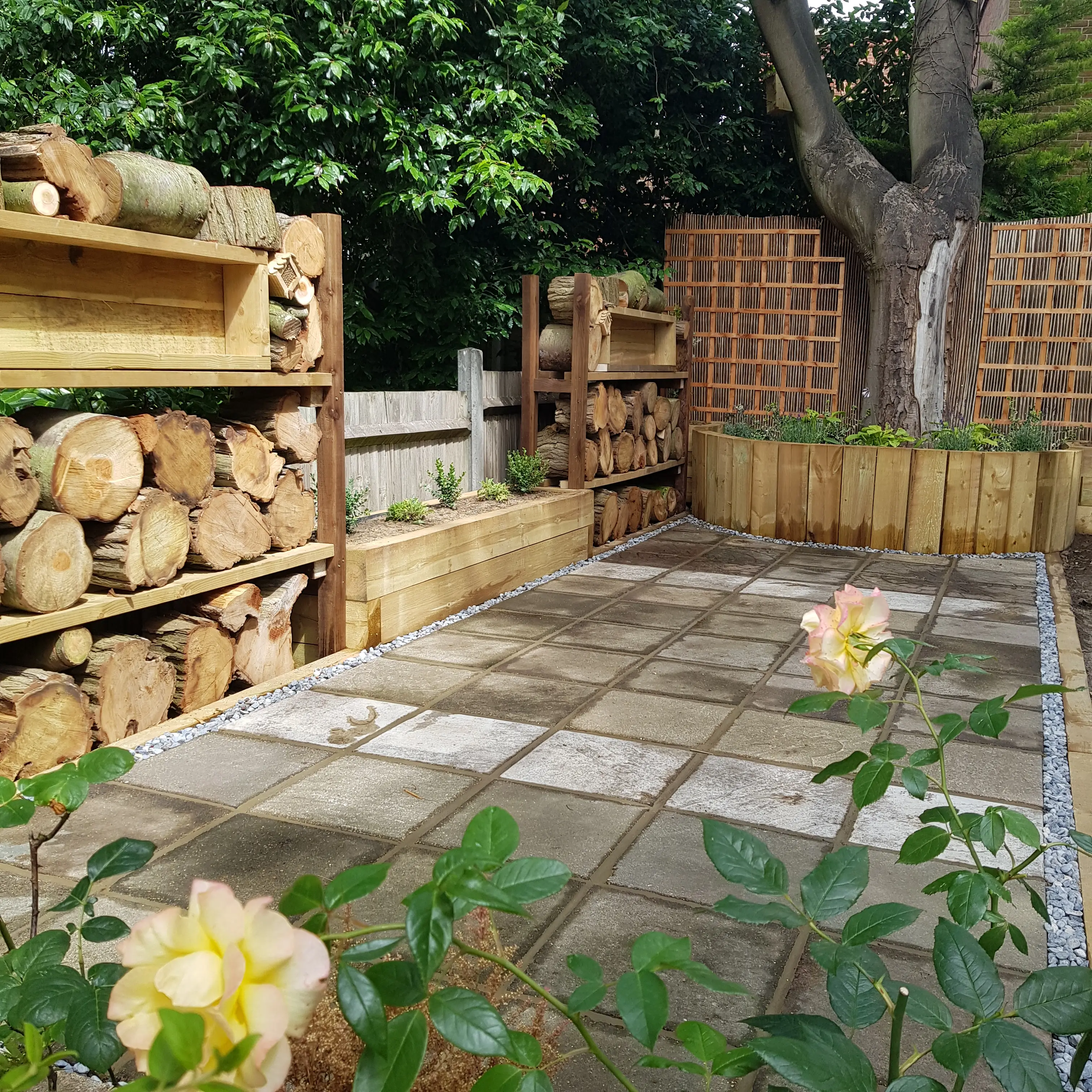 A peaceful garden scene with stacked wooden logs, paving stones, and blooming yellow flowers.