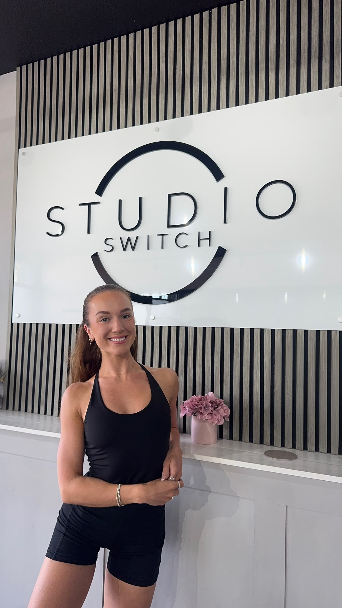 Kirby, a Studio Switch Pilates instructor, standing at the reception counter beneath the Studio Switch sign.