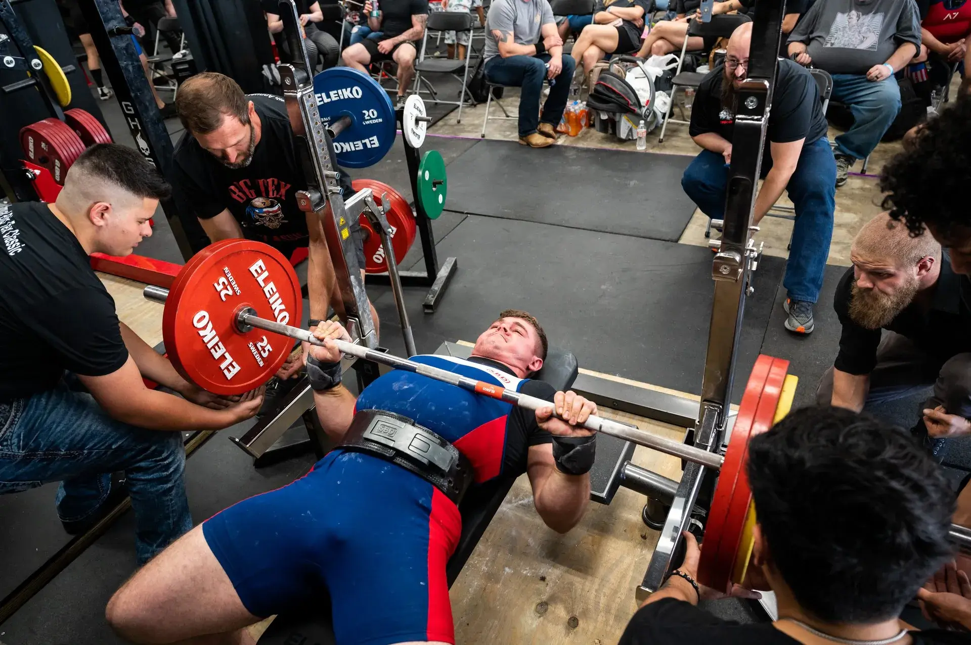 Athlete performing a heavy bench press at a powerlifting competition surrounded by spotters