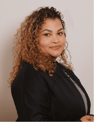 Retrato de uma mulher com cabelos cacheados castanho-claros, vestindo jaqueta preta sobre blusa clara em fundo neutro