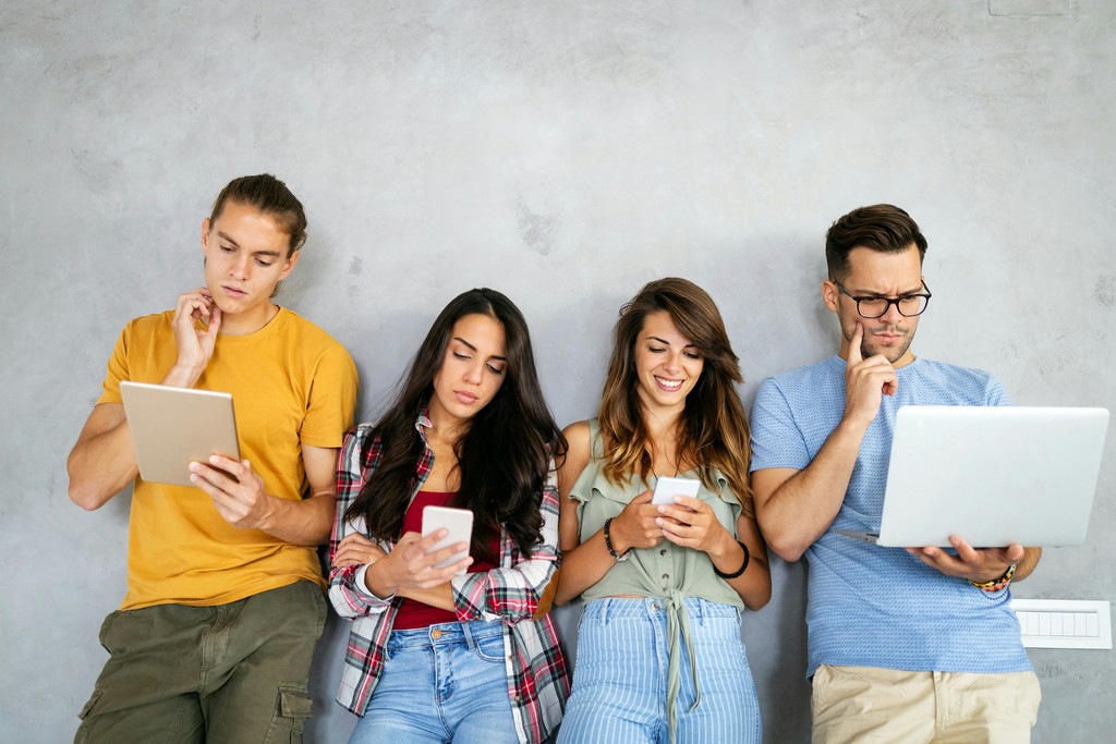 A group of four young adults stand against a gray wall, each engrossed in their digital devices—tablets, smartphones, and a laptop—illustrating the pervasive role of technology and social media in modern life.