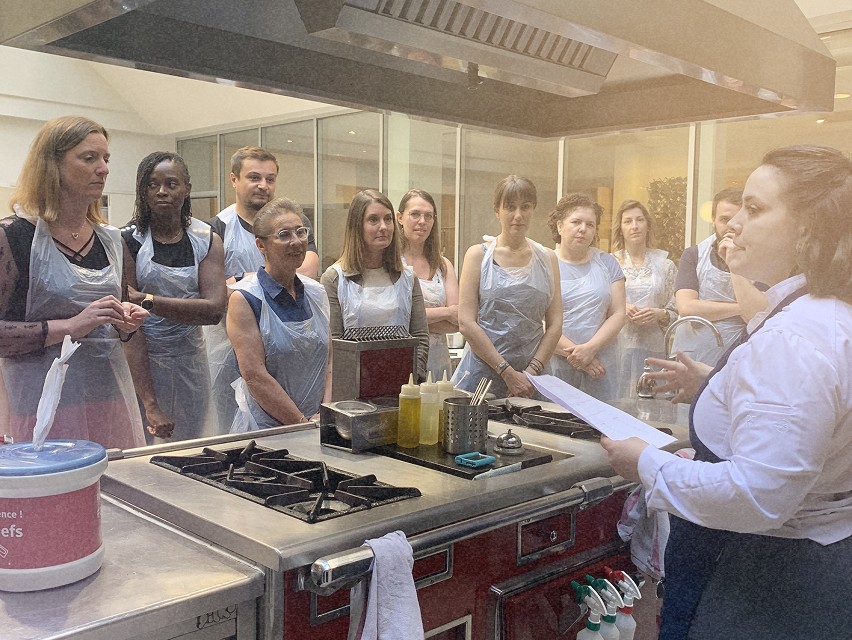 Groupe de personnes portant des tabliers écoutant une chef dans une cuisine moderne avec des ustensiles en préparation.
