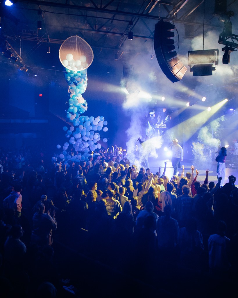 Three young women stand together outdoors, smiling and holding drinks, with cars and trees in the background.