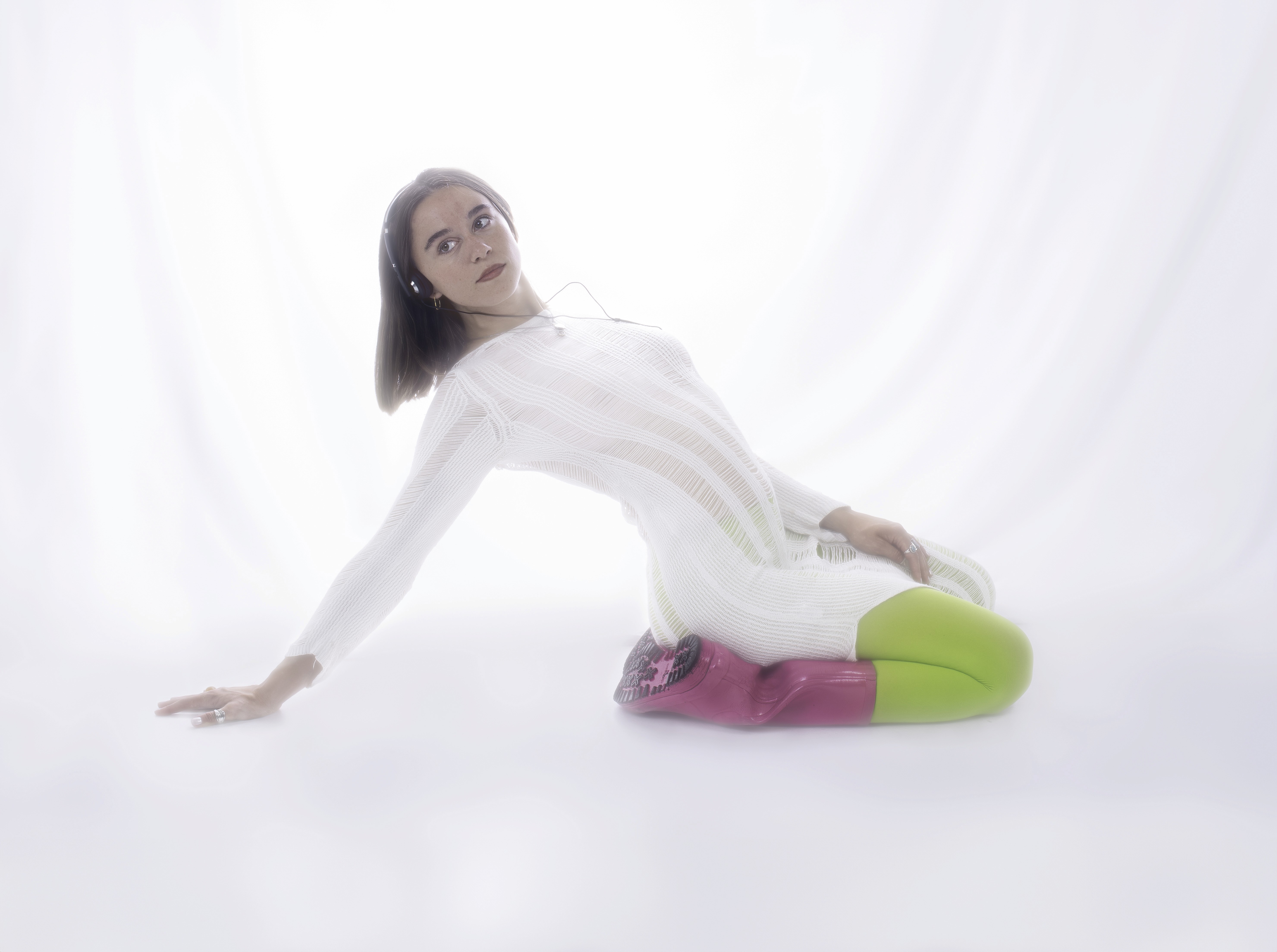 Anna Róshildur, the artist behind Róshildur, poses in a soft, minimal studio setting, wearing a sheer white dress with neon green tights and pink boots, leaning back against a bright, ethereal white backdrop. Photo: Melkorka Embla 