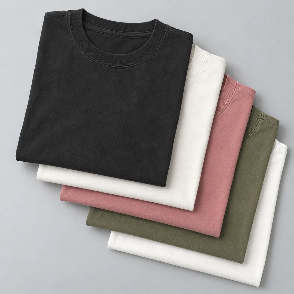 Five folded heavy tees stacked: black, white, rose, olive and white