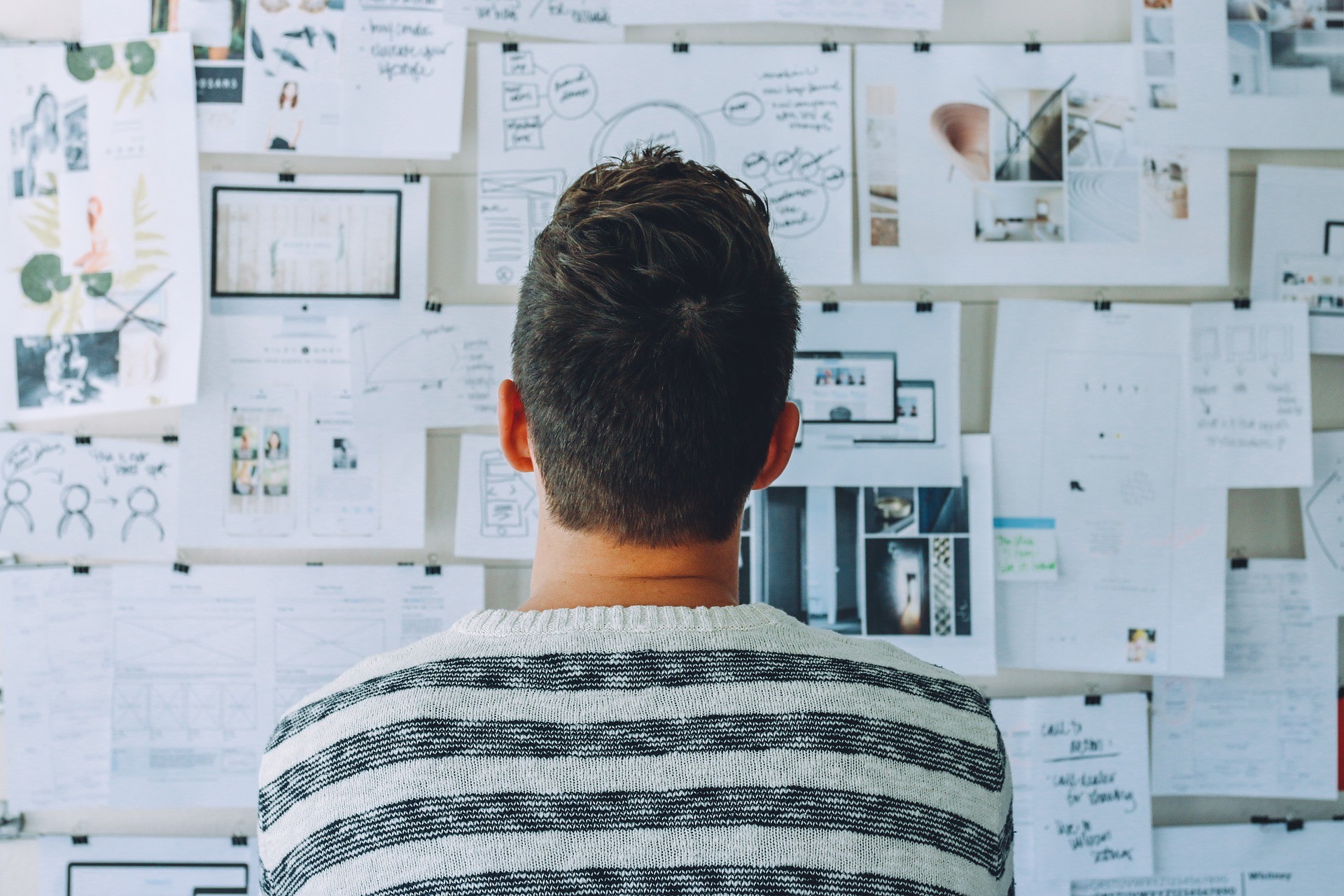A person in a striped sweater stands with their back to us, intently studying a wall covered in various design notes and.