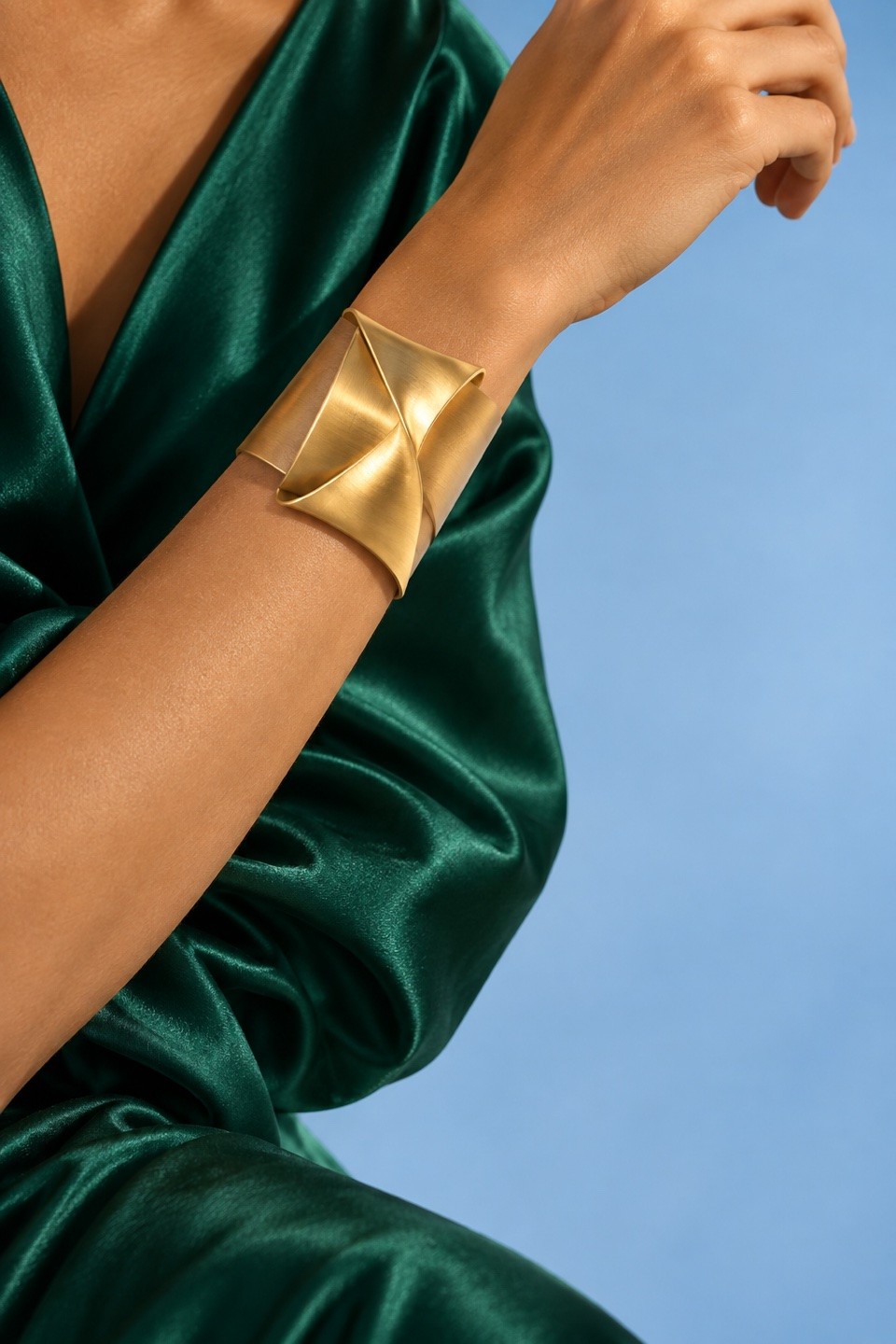 Sculptural folded gold cuff worn against emerald silk and a powder-blue backdrop