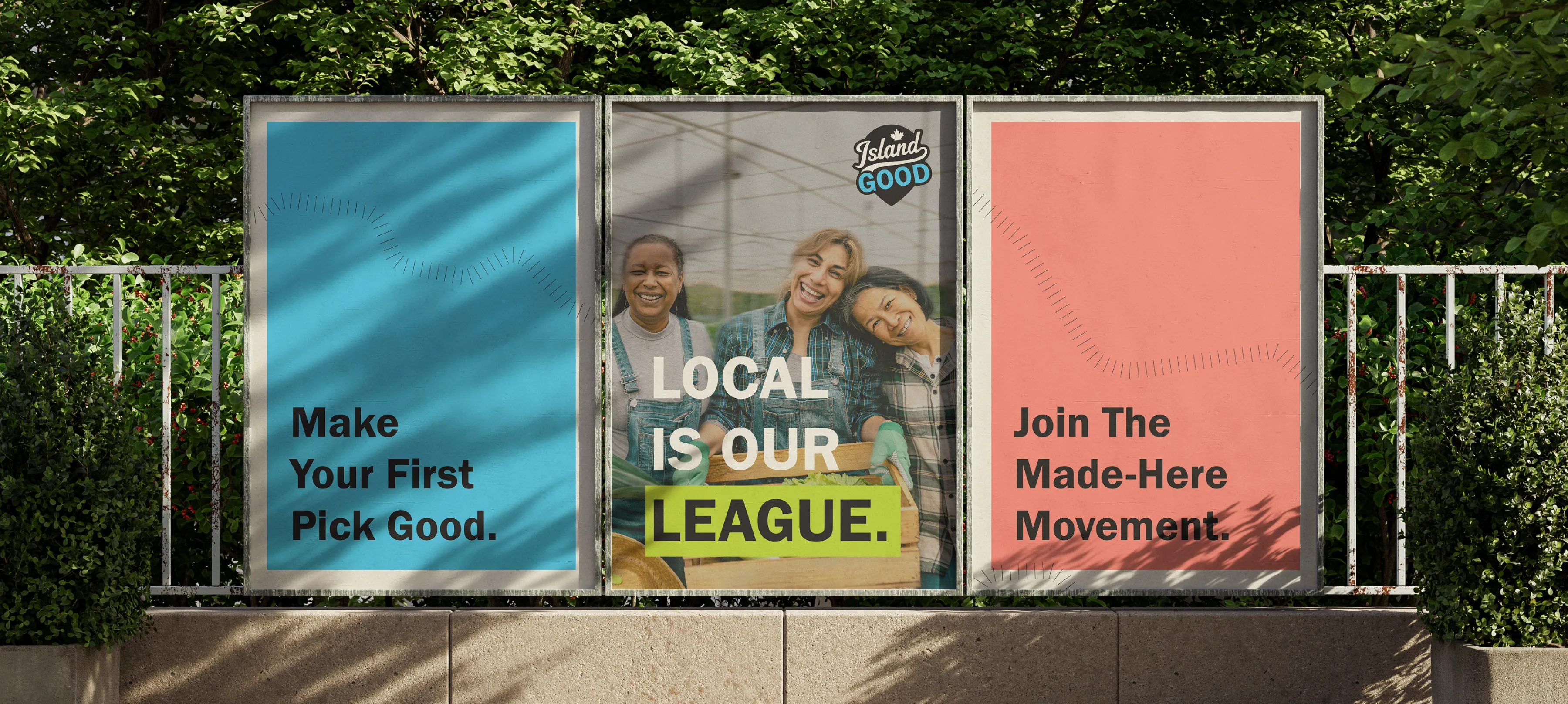 Three posters in frames outside in front of a green space and a fence. The first poster says "Make your first pick good" on a sky blue background. The second says "Local Is Our League" with league highlighted with a lime green box, 3 women huddled up in the background, and the Island Good logo in the top right corner. The third says "Join the Made-Here Movement" on a coral background.