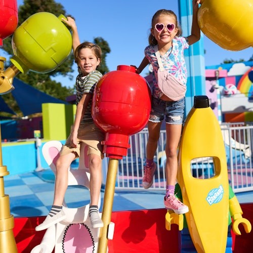 Two children smiling and playing on colorful playground equipment, with oversized red, green, and yellow fruit sculptures around them.