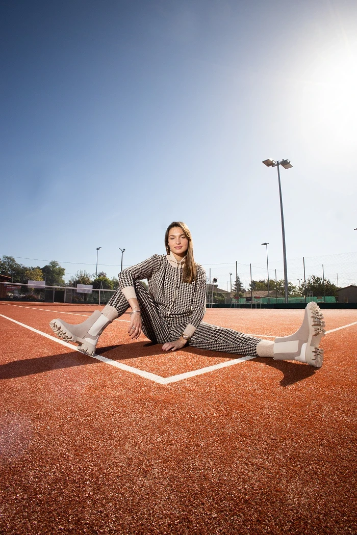 Une femme avec un ensemble à motifs gris. Elle est assise au sol sur un court de tennis.