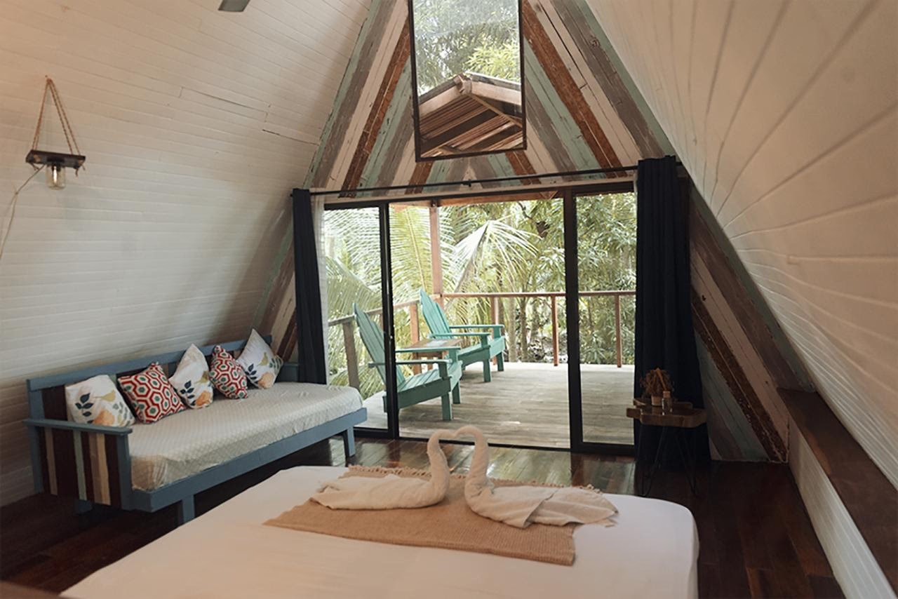 Interior of an A-frame cabin with a white bed, patterned sofa, and glass doors leading to a wooden balcony.