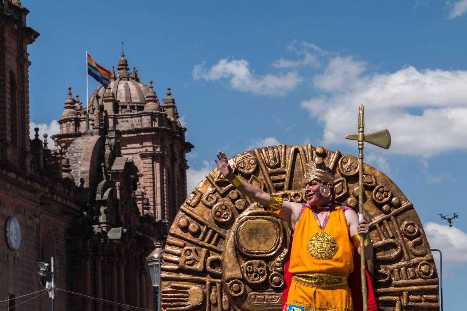 The Sun God, Inti Raymi, stands in front of a semi-circular sculpture waving at the crowds as part of a procession in Cusco to celebrate the winter solstice. In the background: a building and flag.