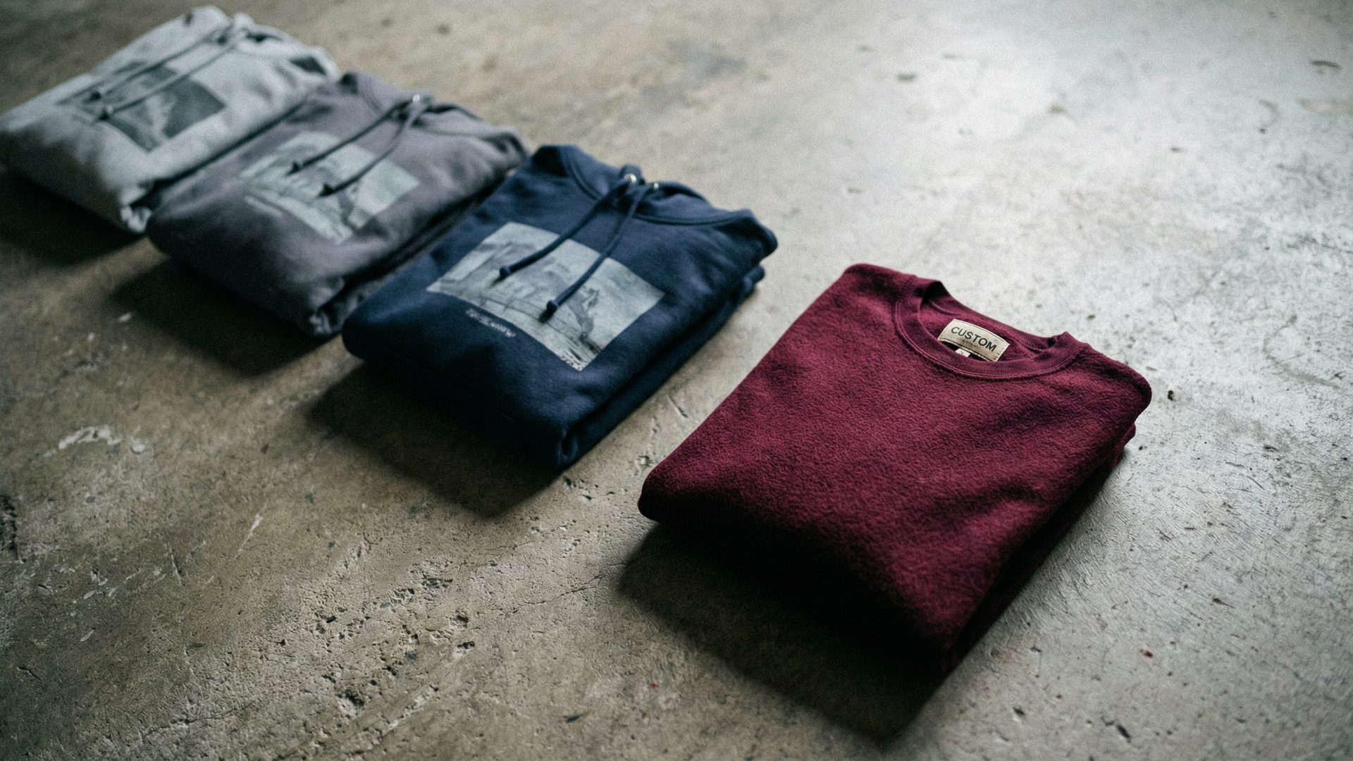 Three identical screen-printed hoodies in grey and navy laid side by side on a concrete surface, contrasted with a single folded custom-cut garment in rich burgundy with a woven label visible at the collar, illustrating the difference between generic blanks and a custom cut-and-sew product.