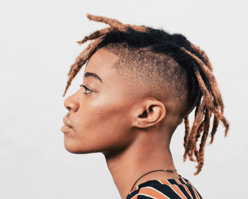 A black person stands against a white wall. The sides of their hair are shaved, with dreadlocks on top. They are in portrait, facing to the left.