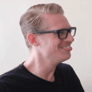 A person with glasses and sunglasses smiling by a wall.