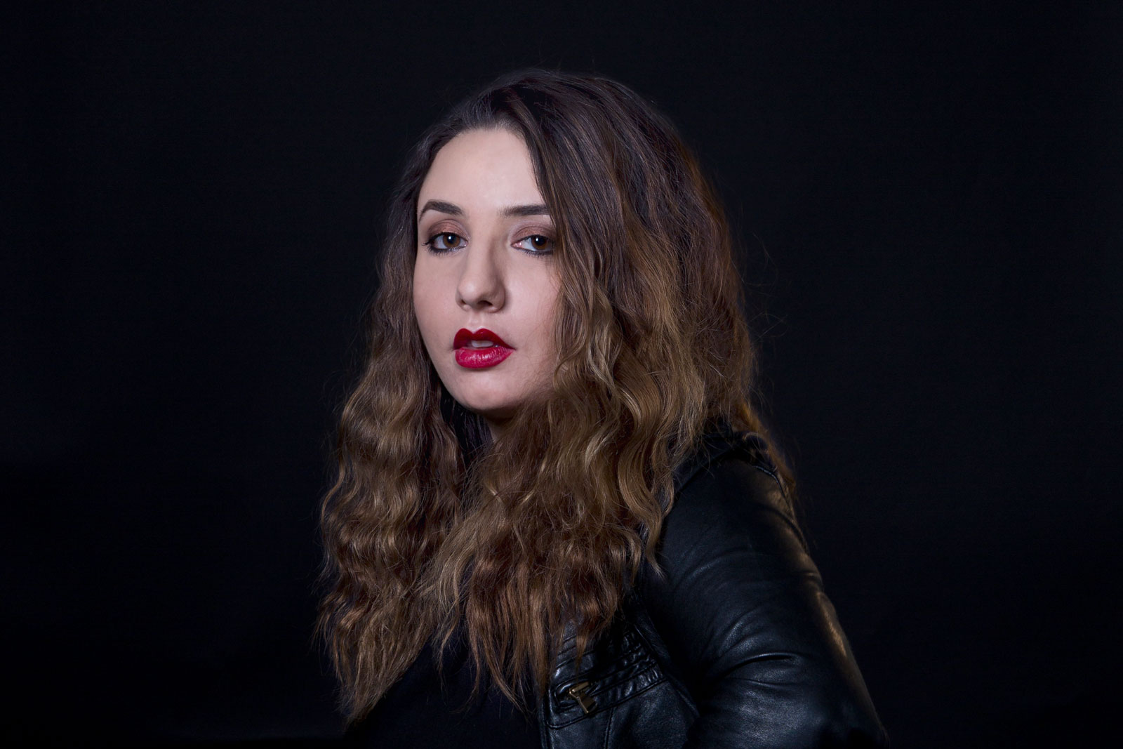 A young woman with long, wavy brown hair and dark eyes stands against a solid black background. She wears a black leather jacket and bright red lipstick. Her gaze is directed slightly to her left. The lighting is focused on her face and upper body.