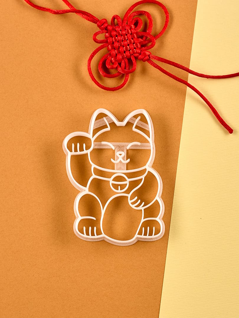 Obraz prezentuje foremkę do ciastek Biocraftlab w kształcie machającego kota, umieszczoną na tle brązowego i żółtego papieru z ozdobnym czerwonym węzłem powyżej.