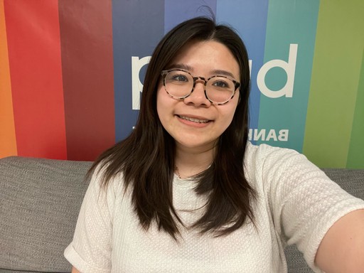 A smiling person with glasses poses for a selfie in front of a colorful background.