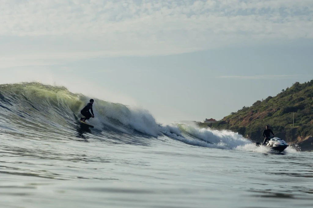 Surfer riding a wave with Jet Ski safety support nearby.