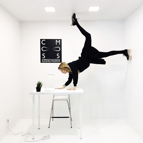 一位女士在纯白色房间的桌子上做倒立，墙上挂着单色幻觉博物馆的标志。