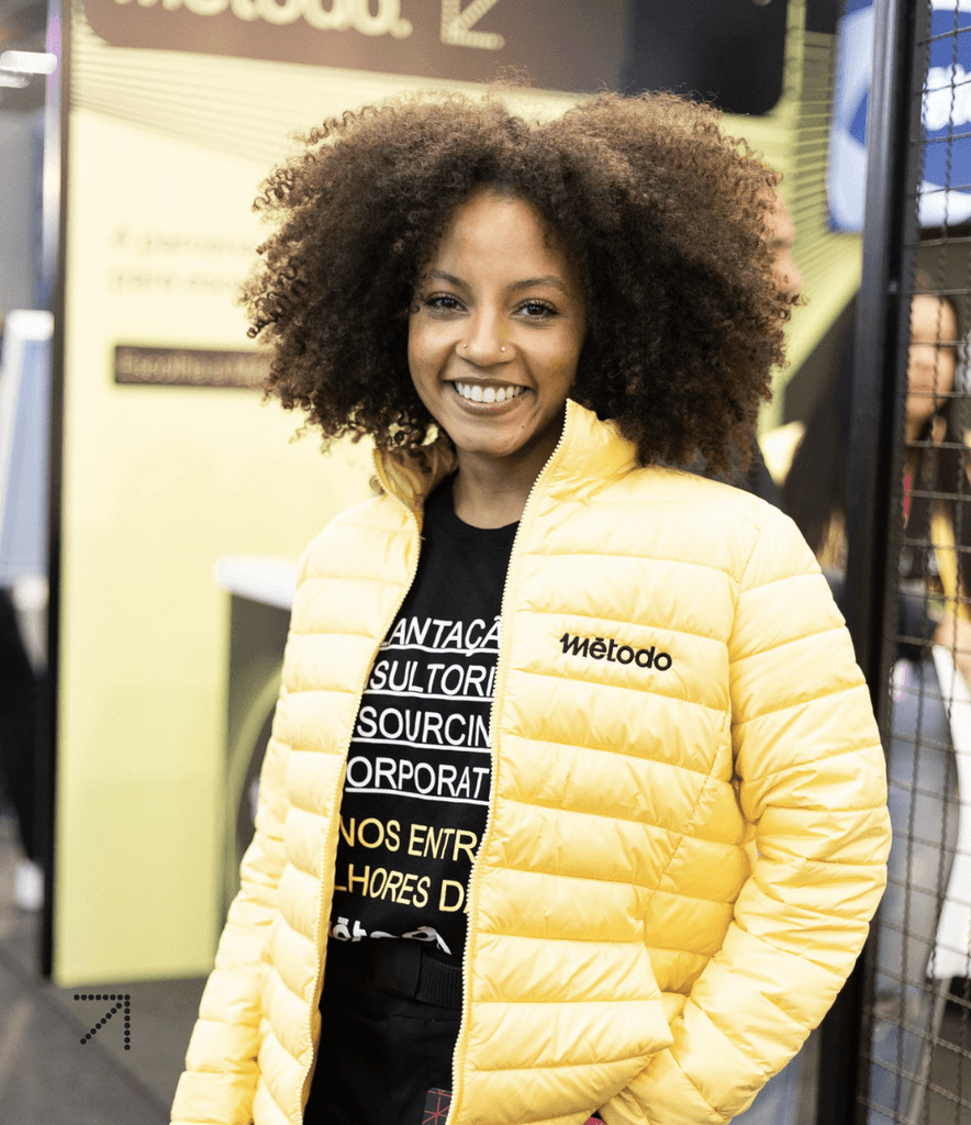 Colaborador Método, vestindo uma jaqueta amarela brilhante e com cabelo cacheado, em pé em um ambiente interno animado.