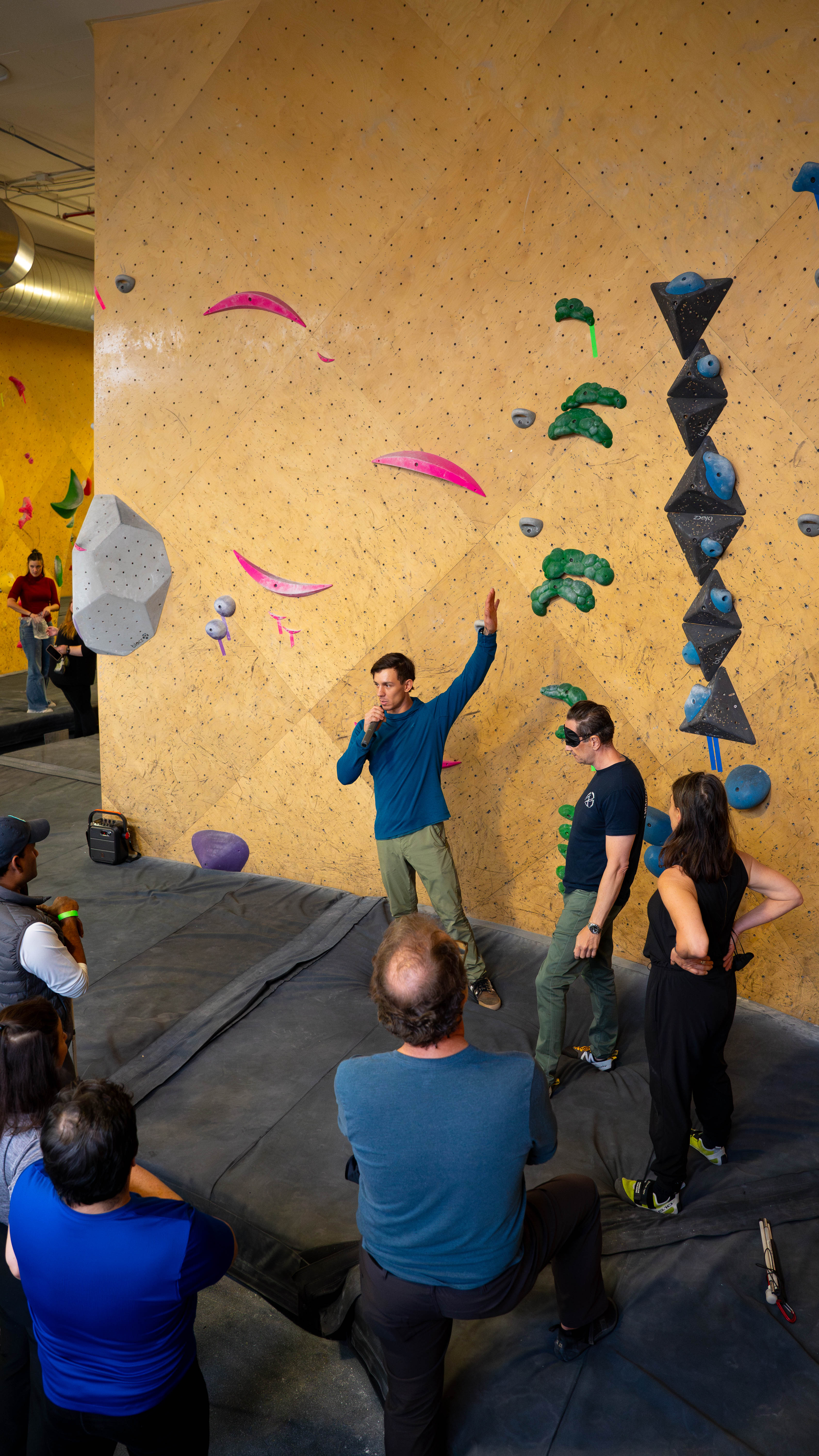 Justin stands in front of a bouldering wall, explaining the concepts of climbing without sight. To his left, a volunteer wears a blindfold. A group can be seen along the edges of the photo