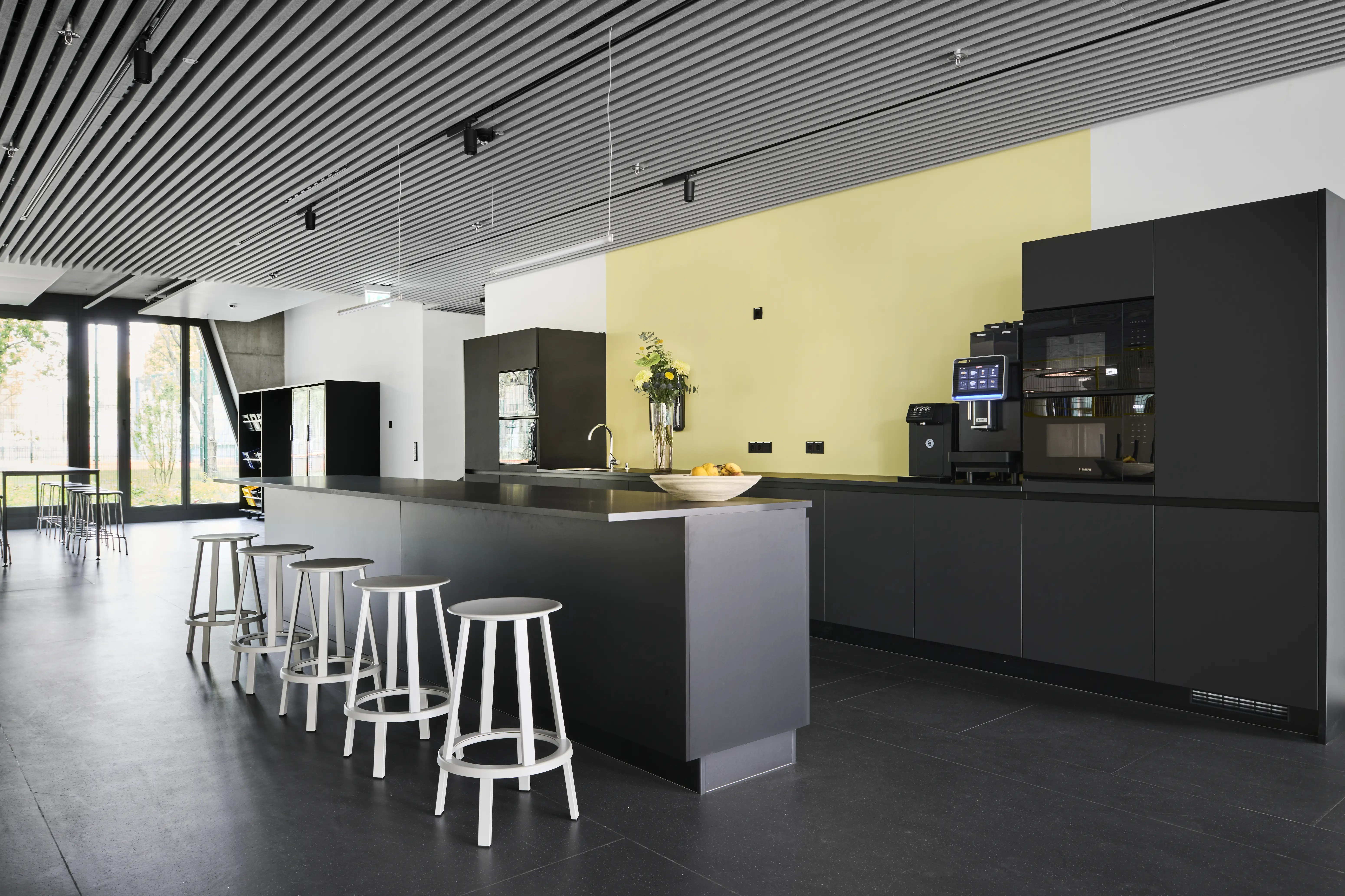 Black kitchen island at ENPAL with white round bar stools, a pale yellow accent wall, built-in coffee machines and a slatted black ceiling with track lights.