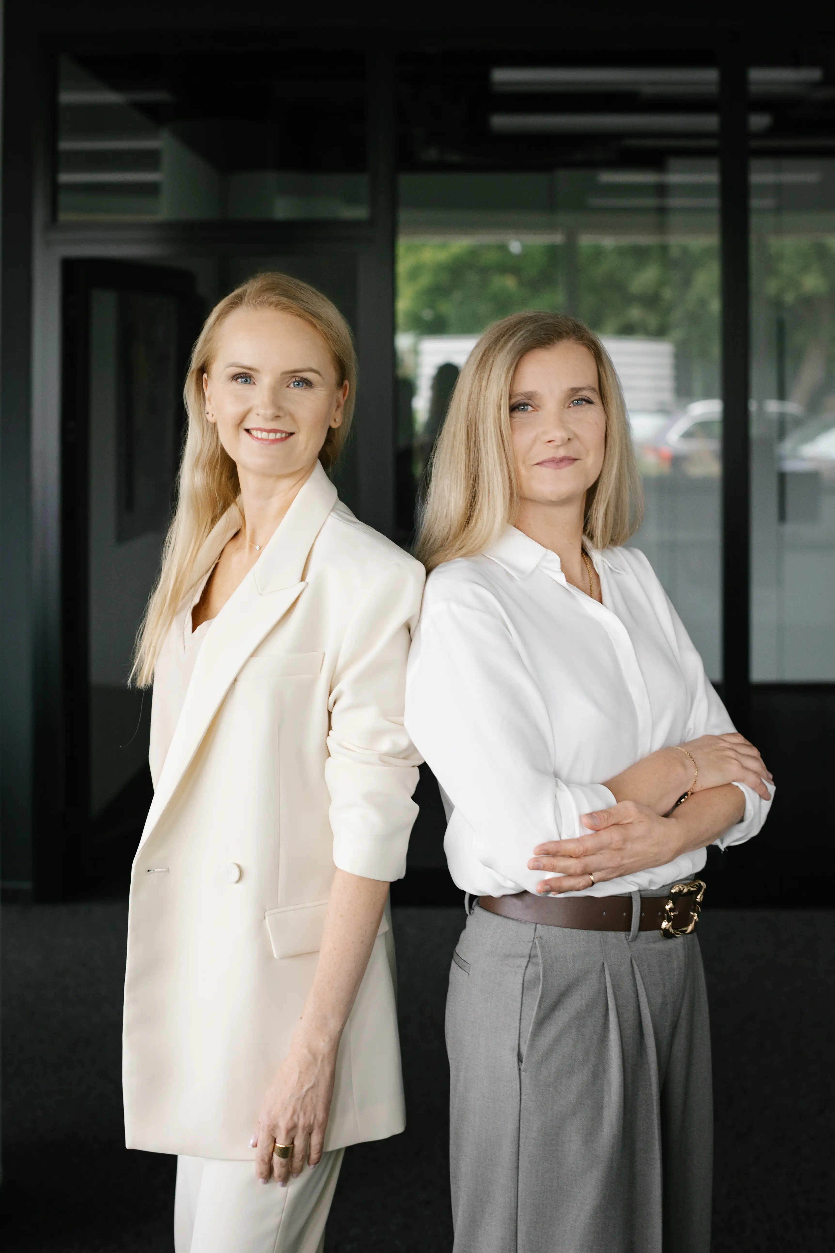 Dwie kobiety w biurowym otoczeniu, stojące obok siebie. Po lewej stronie ubrana w jasny garnitur z długimi włosami, po prawej w białej koszuli i szarych spodniach. Tło biura z dużymi oknami.