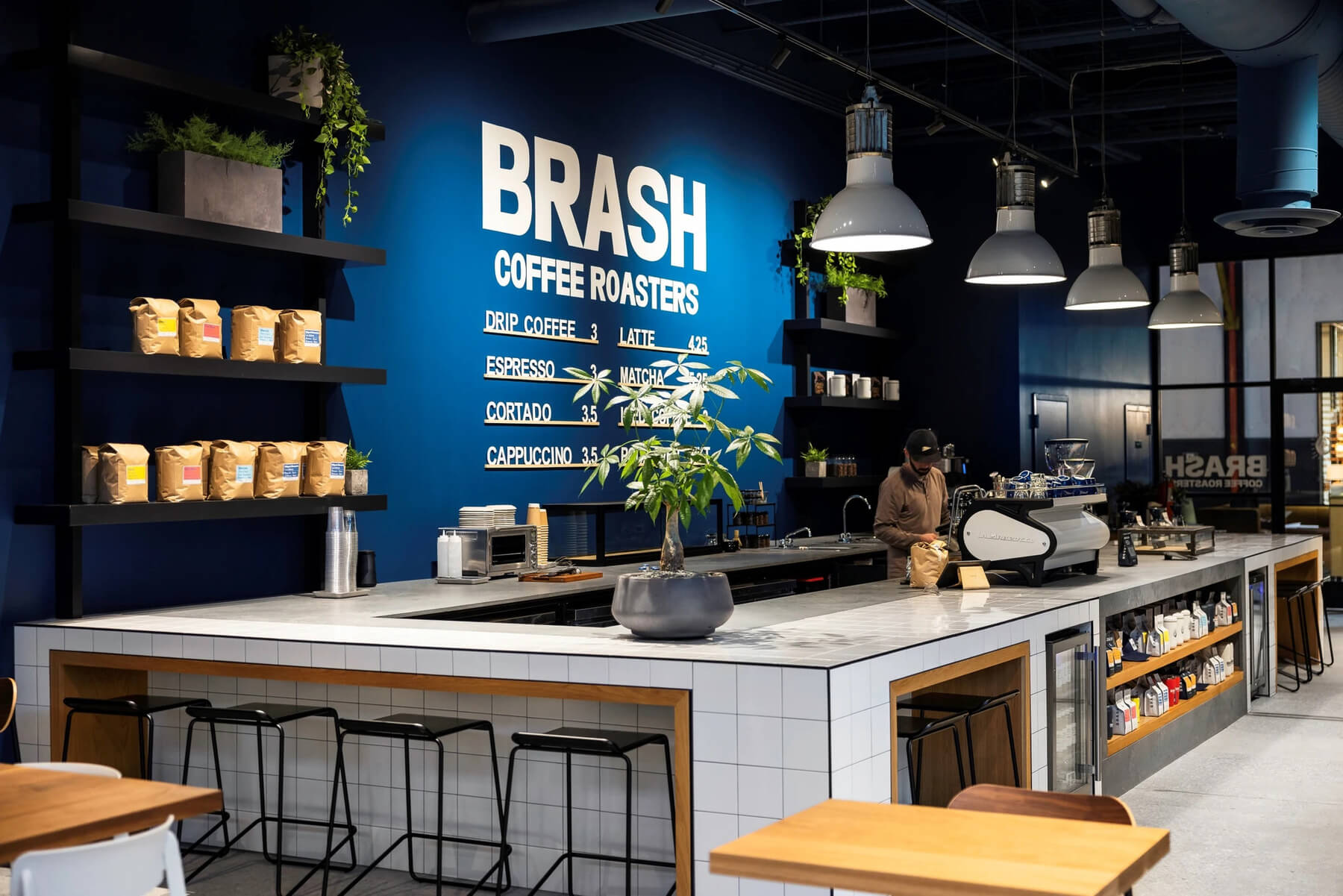 Interior of Brash Coffee Roasters featuring a white tile service bar, menu wall, espresso machine, industrial pendant lighting, and packaged coffee bean shelves.