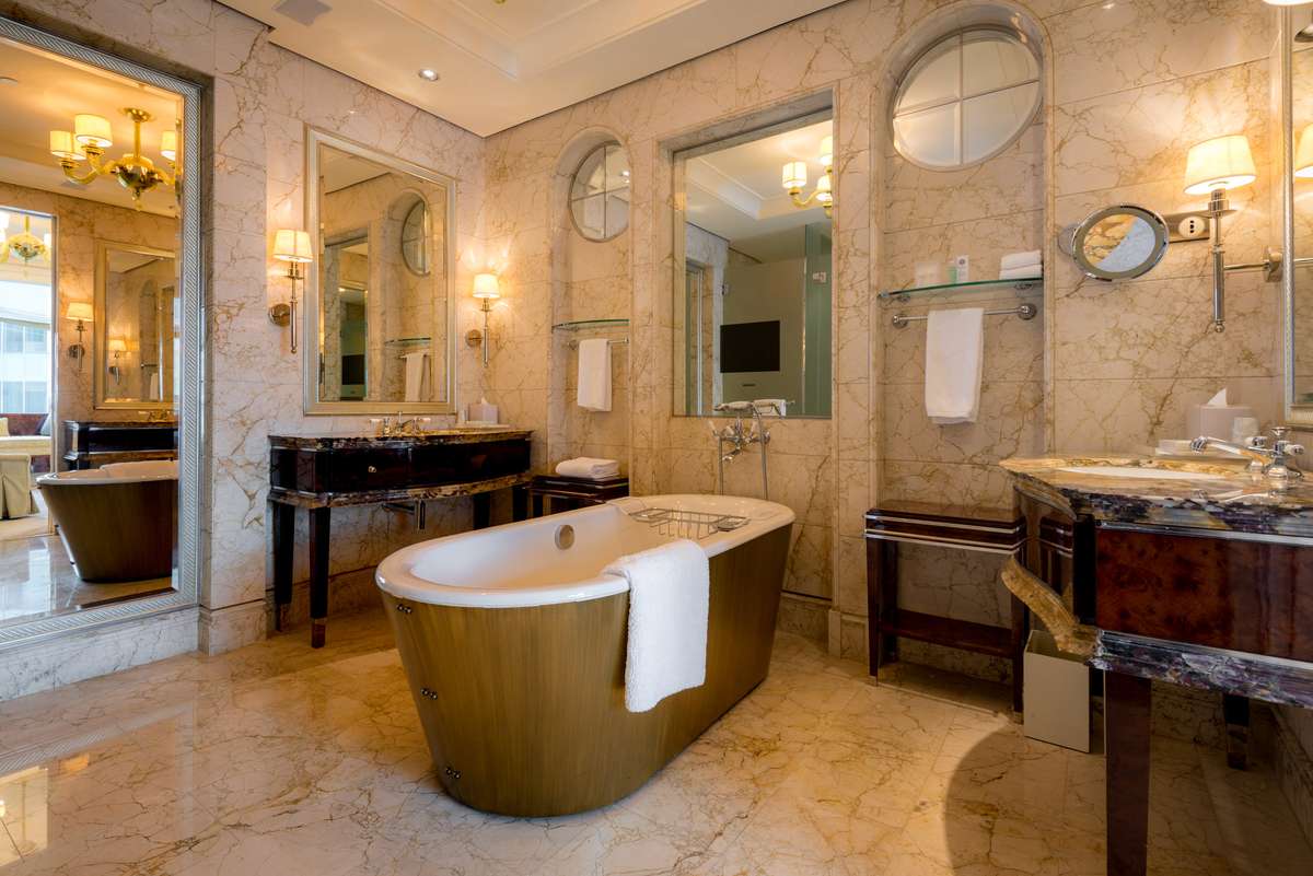 A marble-tiled bathroom features a freestanding copper tub, two dark wood vanities with white sinks, and large mirrors under warm lighting.