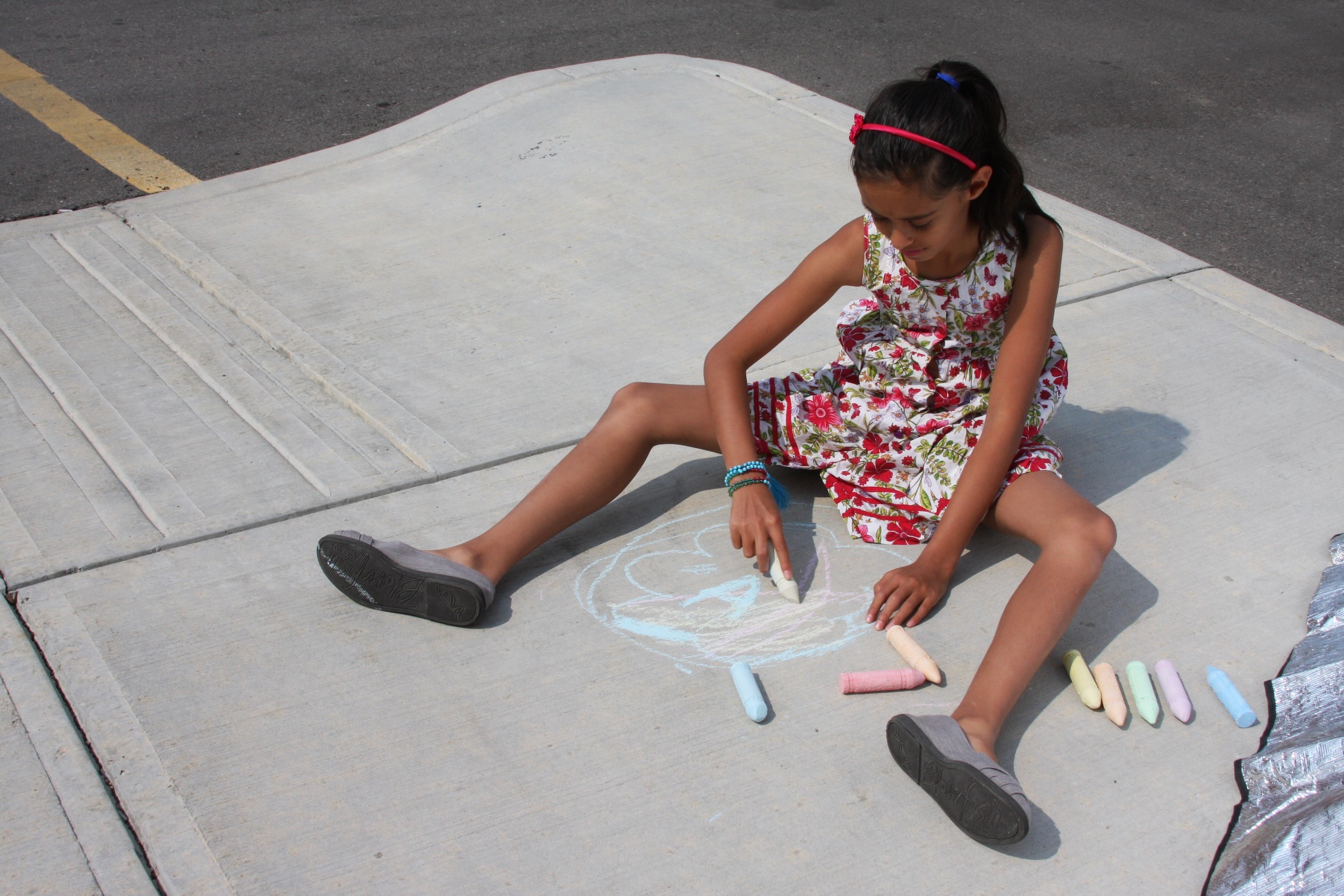 A girl in a flowered dress sits on the pavement drawing in coloured chalk.