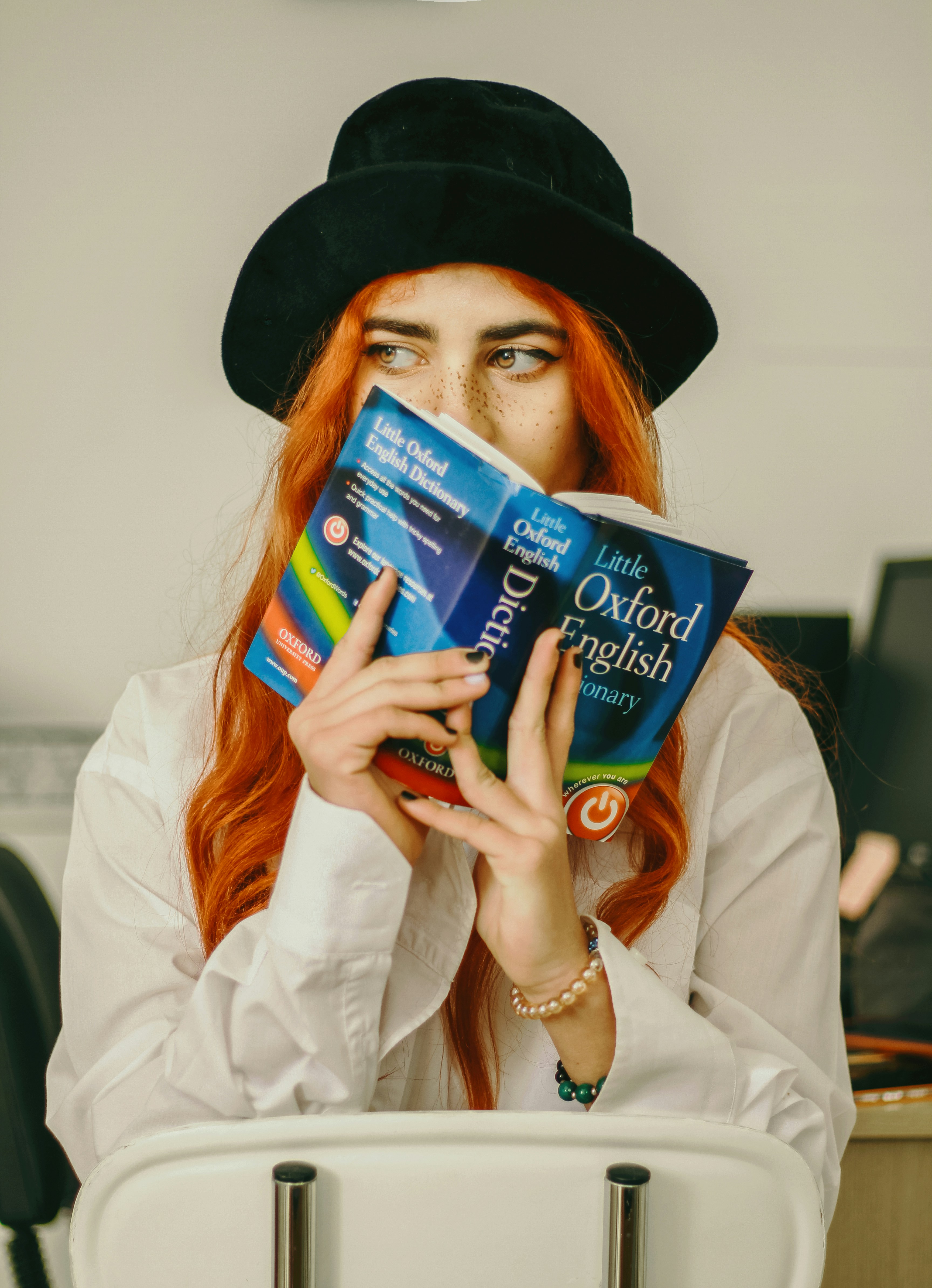 Female with red hair, wearing a black hat reading an Oxford Dictionary
