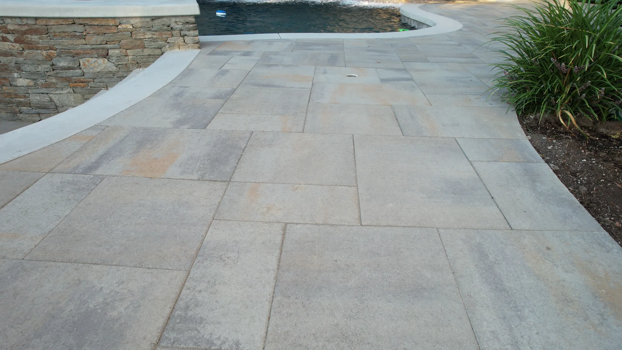 Stone paving curves around a dark swimming pool beside a planted border.