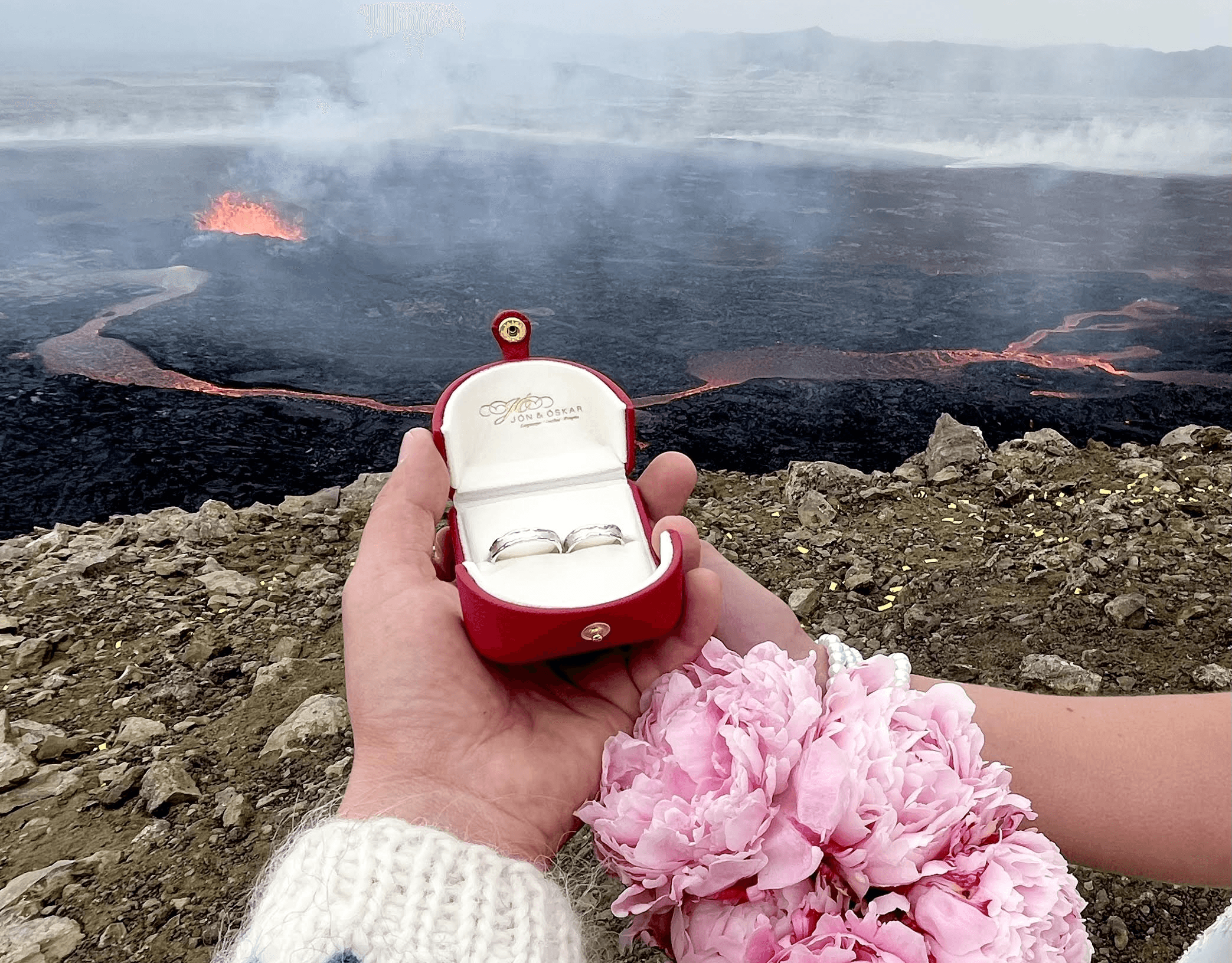 A hand holds an open ring box with the Northern Lights in the sky and rocky terrain in the background, symbolizing a proposal in Iceland.