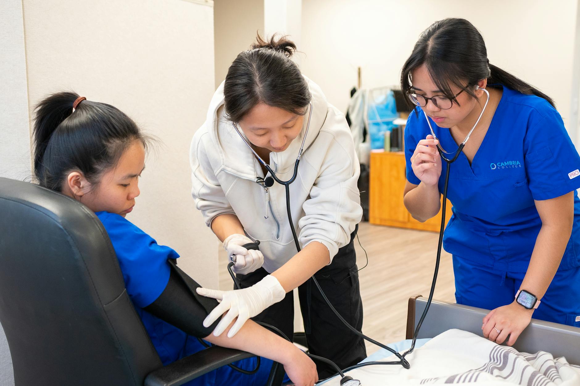 Students in blue scrubs practicing blood pressure readings on a medical mannequin in a clinical lab setting.