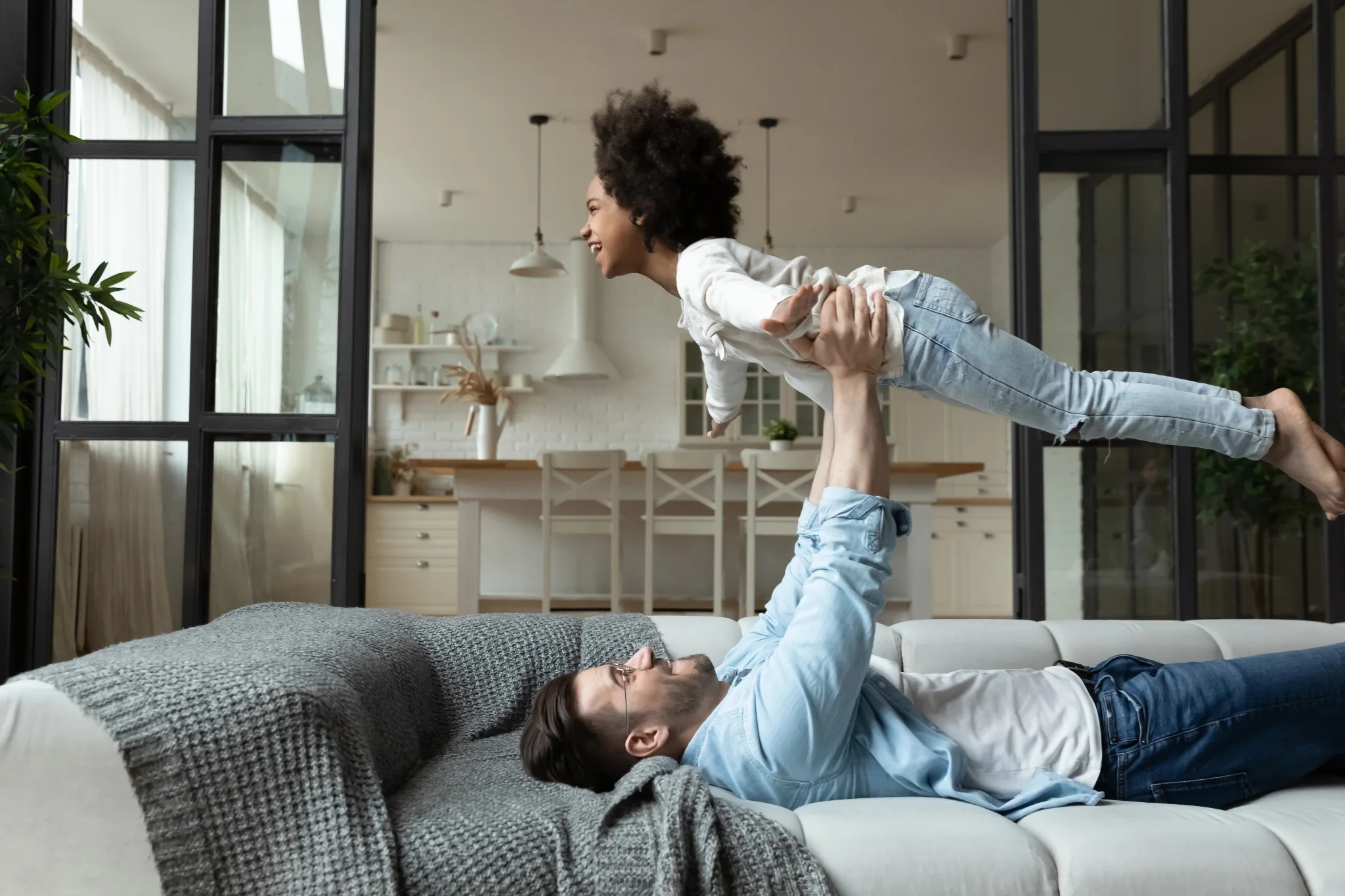 Man laying on a couch, holding a child in the air, kids' hands both up in the air.
