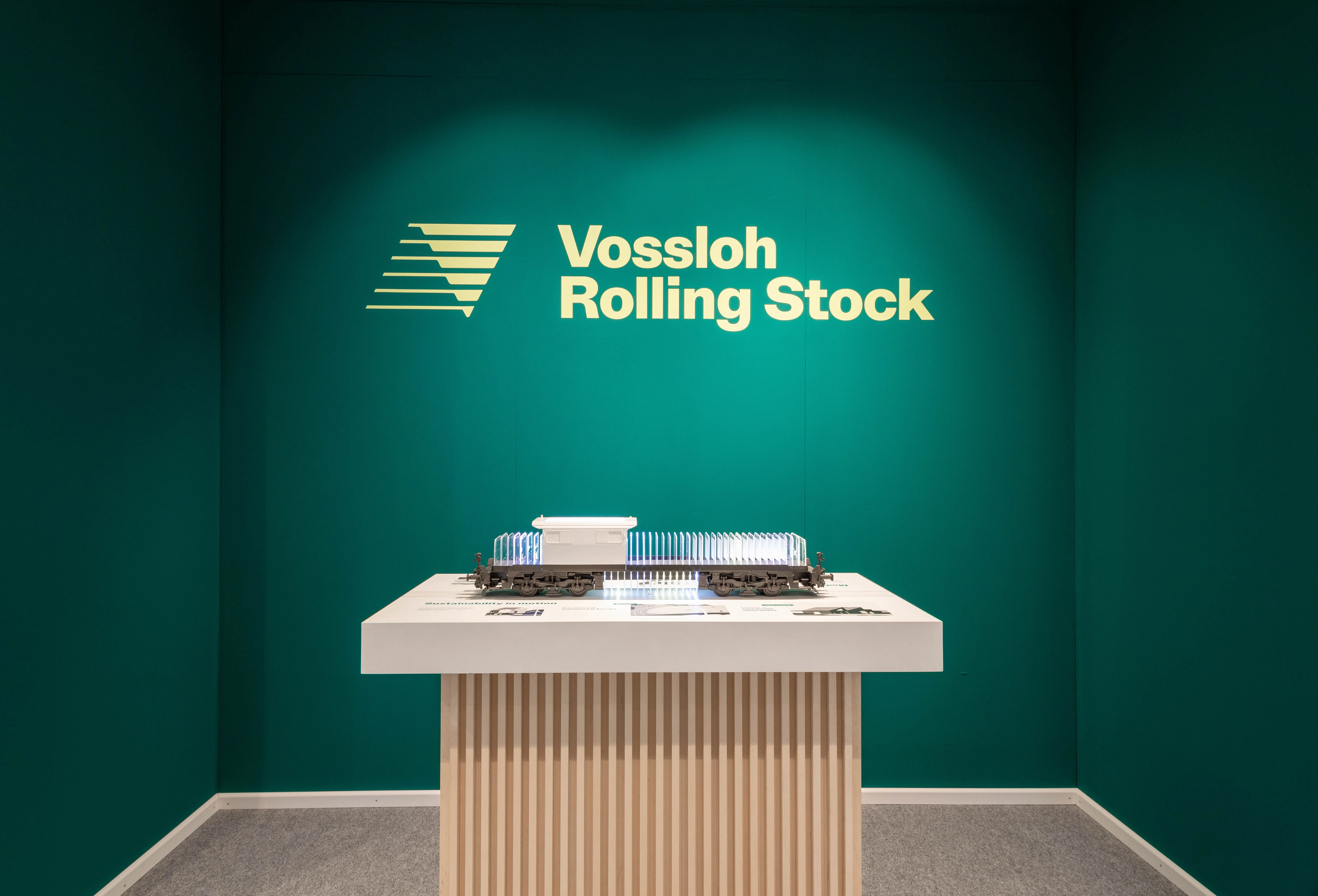 Green presentation niche with yellow Vossloh Rolling Stock logo and locomotive model on a slatted-wood plinth.