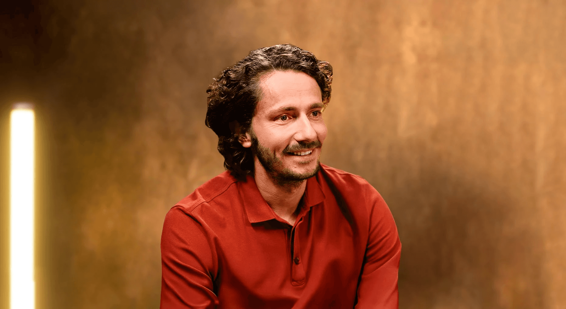 Portrait professionnel et souriant de Guillaume Pley, fondateur du média digital Legend, dans son studio d'enregistrement. Il est vêtu d'un polo rouge terre cuite. Le sujet regarde vers la droite, laissant un large espace négatif à droite de l'image. L'arrière-plan est un mur texturé, flou, dans des tons chauds (or, bronze, marron), éclairé par une colonne lumineuse verticale à l'extrême gauche, créant une ambiance de studio d'interview et de podcast haut de gamme. 