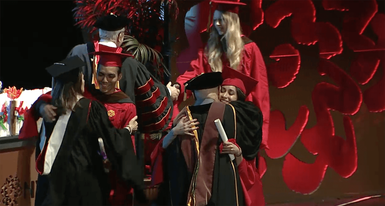 A photo of a couple of grad students and teachers, dressed in SVA color robes, that are hugging and congratulating each other.