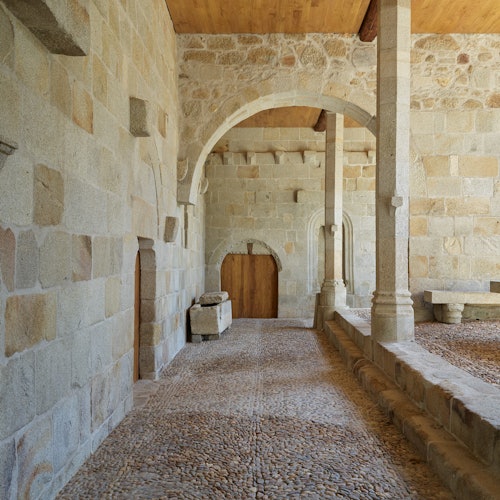 一条石质走廊，两旁有拱形门道和柱子，还有一扇木门。地板由鹅卵石铺成。