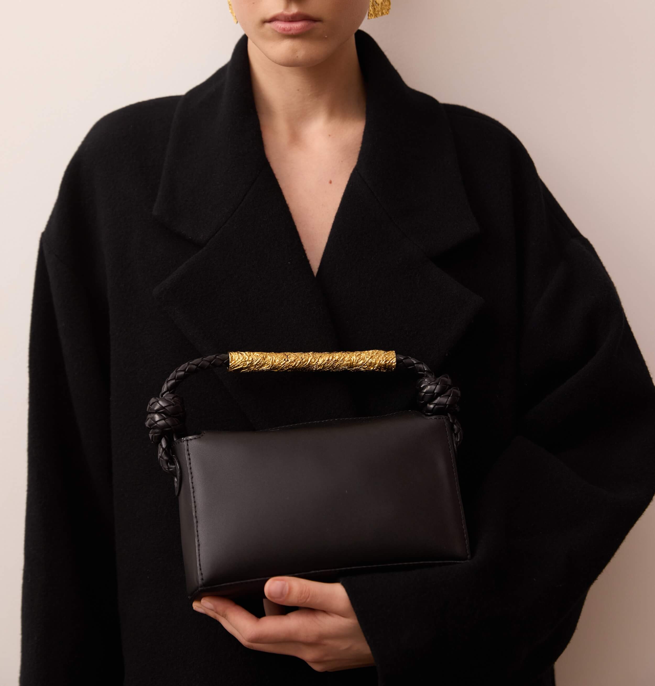 Close-up of a person in a dark coat holding a large black leather tote with brown straps, with a woven basket bag slung over the shoulder.
