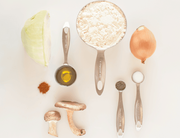 The image shows a white background with various food items and measuring spoons arranged on it. On the left side, there is a halved light green cucumber or zucchini. Next to it is a small pile of brown spice, likely cumin or coriander. Near the center, there is a small pile of yellow liquid, possibly olive oil. On the right side, there is a whole light brown onion, a whole light brown mushroom, and a small pile of white granulated sugar. Two measuring spoons are present: one larger spoon with measurement markings and a smaller spoon with measurement markings and text reading “1 Tbsp 1.25 tsp”.