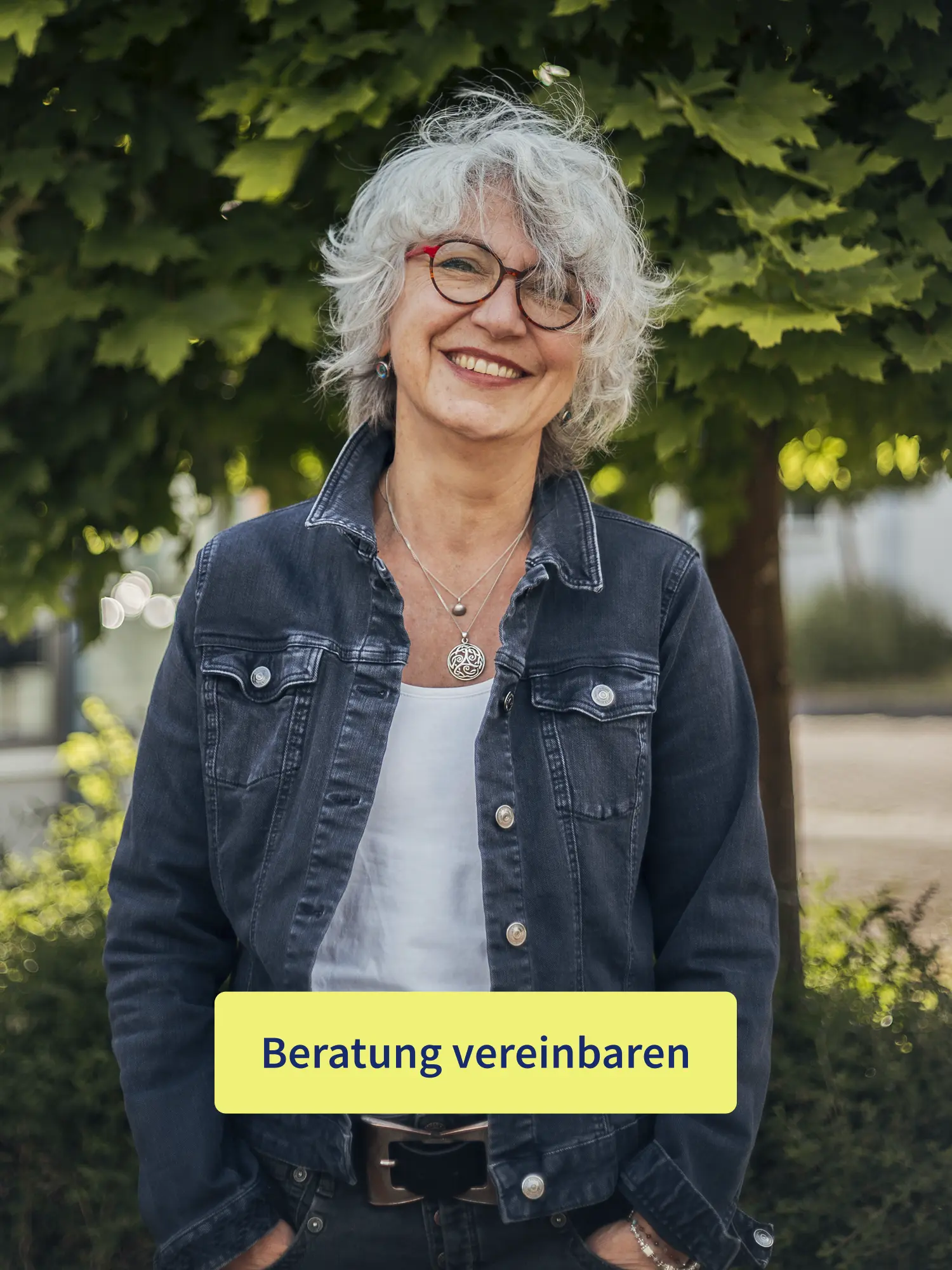Porträt einer lächelnden Frau mit grauen Locken und roter Brille im Garten mit Button „Beratung vereinbaren“.