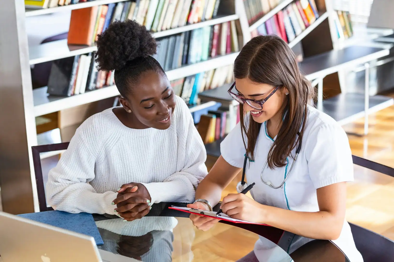 A nurse with a stethoscope reviews clipboard paperwork with a seated patient in a library setting.
