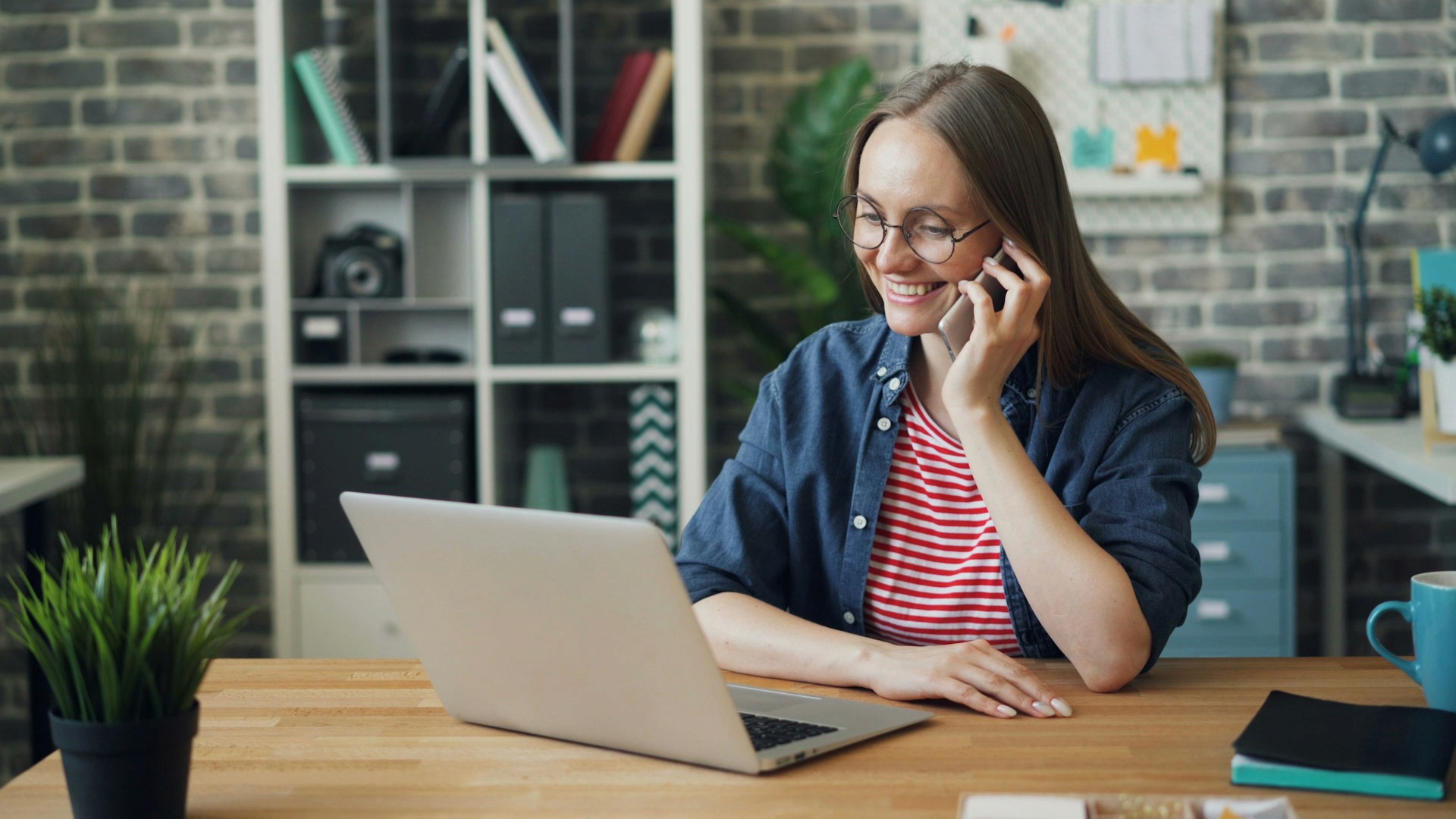 Smiling woman in glasses talking on a smartphone while working on a laptop at a desk.