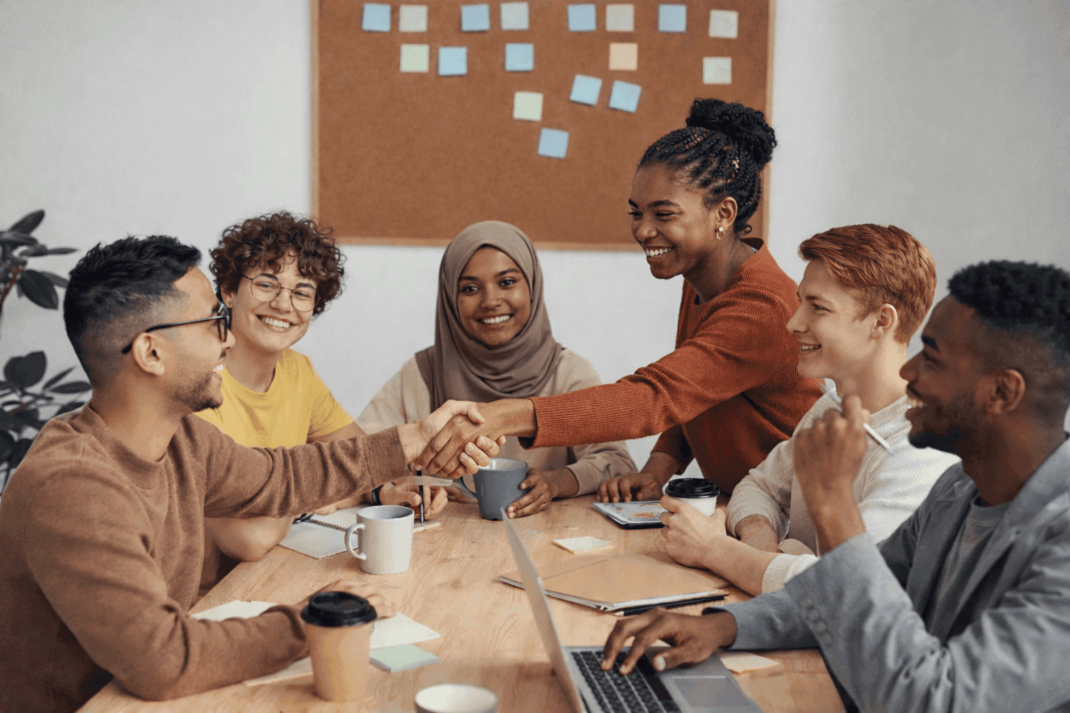 A diverse group of six people, including men and women of various ethnicities, are gathered around a wooden table in a collaborative office setting, with two individuals in the foreground shaking hands, surrounded by laptops, coffee cups, and colorful sticky notes on the wall in the background.